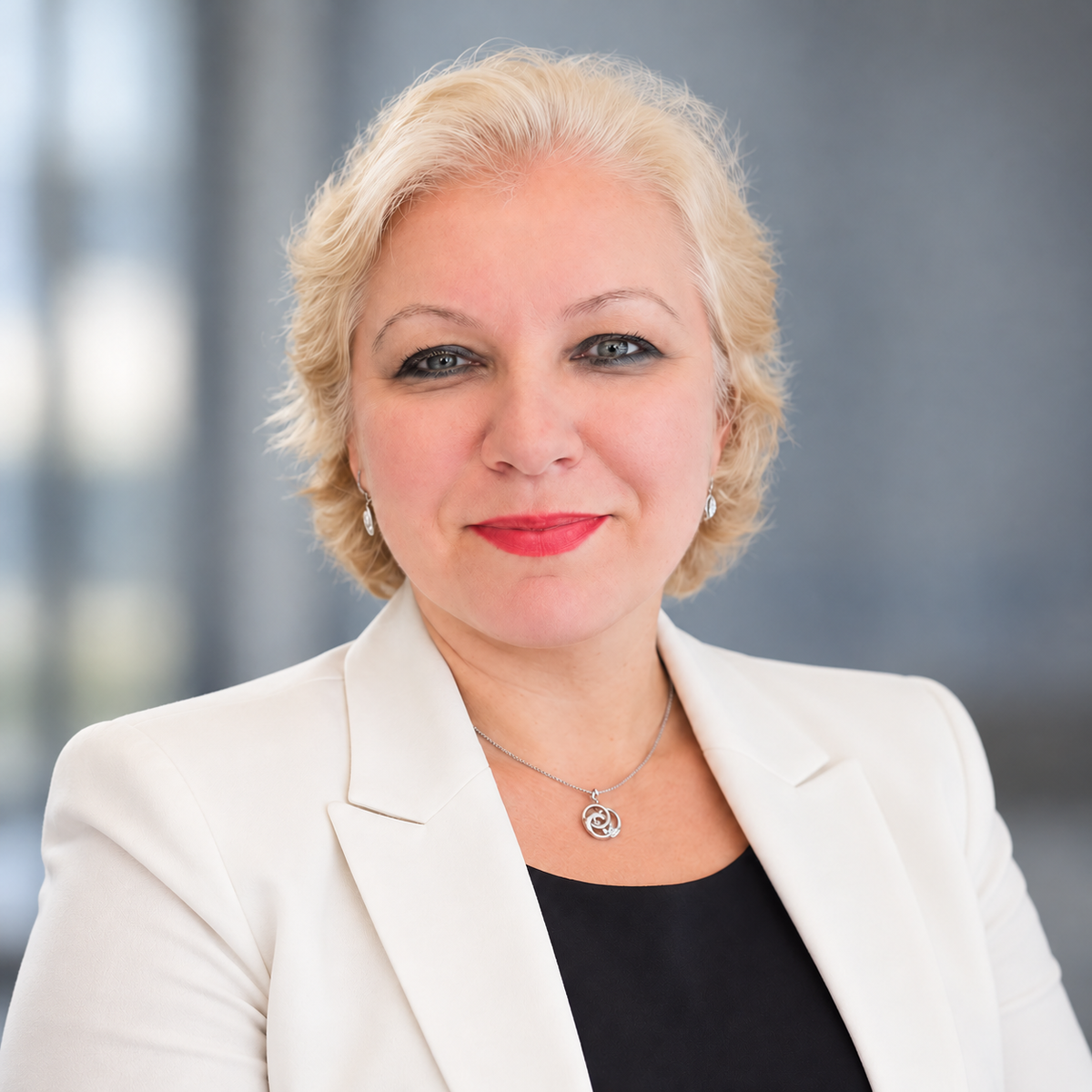 A professional portrait of a blonde woman with blue eyes, wearing a white blazer over a black top, with a silver pendant necklace. The background is a soft, blurred gray office setting.